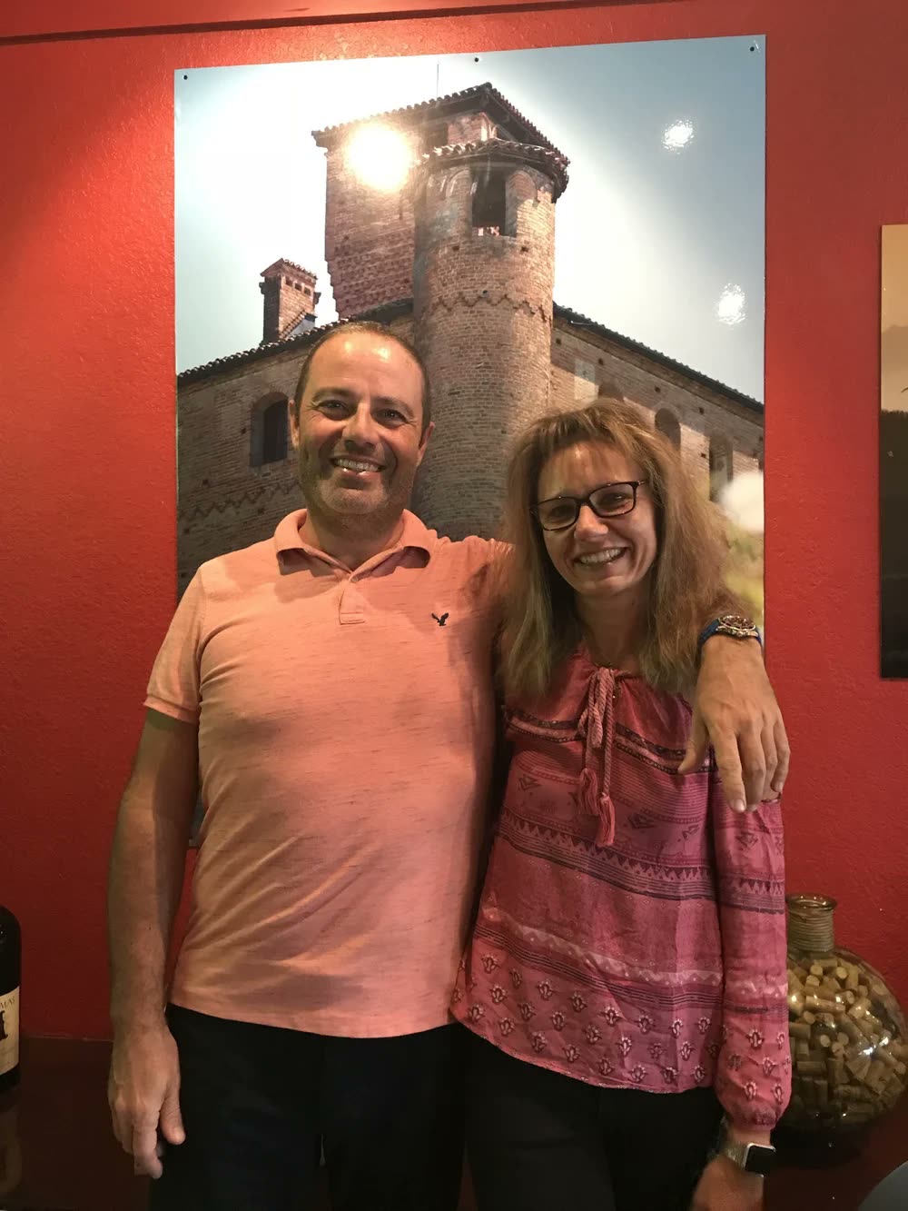 Franco and Mara smiling in front of a photo of a Piedmontese castle on the restaurant's red wall